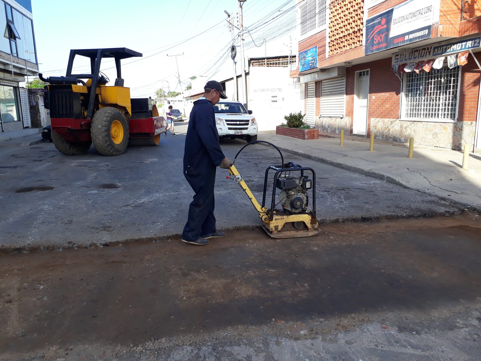 Operario compactando con rana vibratoria junto a vibrocompactador en vialidad urbana